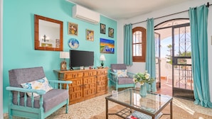 Un salón con paredes verde azulado, un mueble de madera, un televisor, dos sillones, una mesa de centro de vidrio y un balcón con cortinas verde azulado.