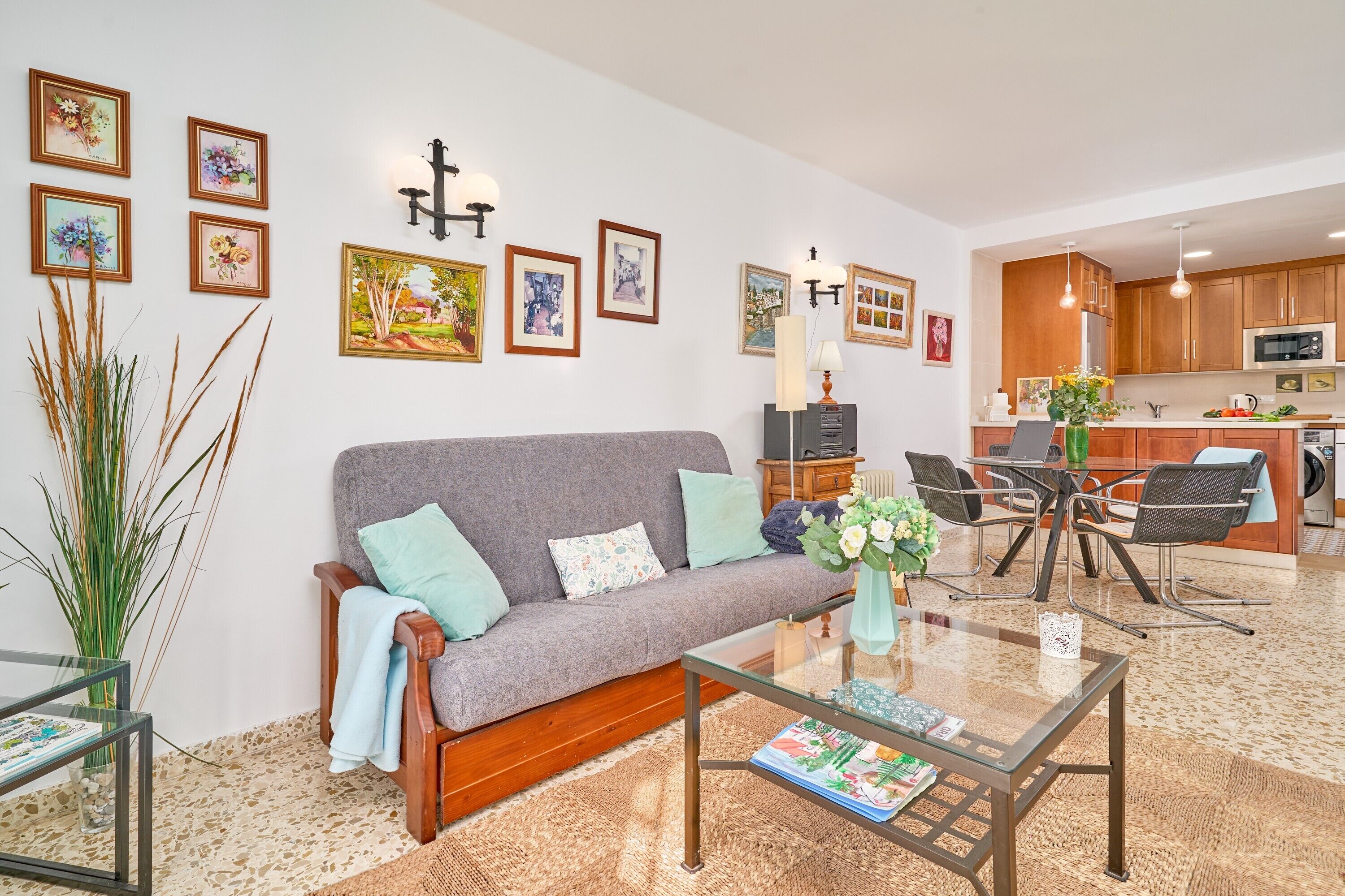 Una sala de estar con un sofá, una mesa de centro y decoración en la pared. Se ve un área de cocina al fondo.
