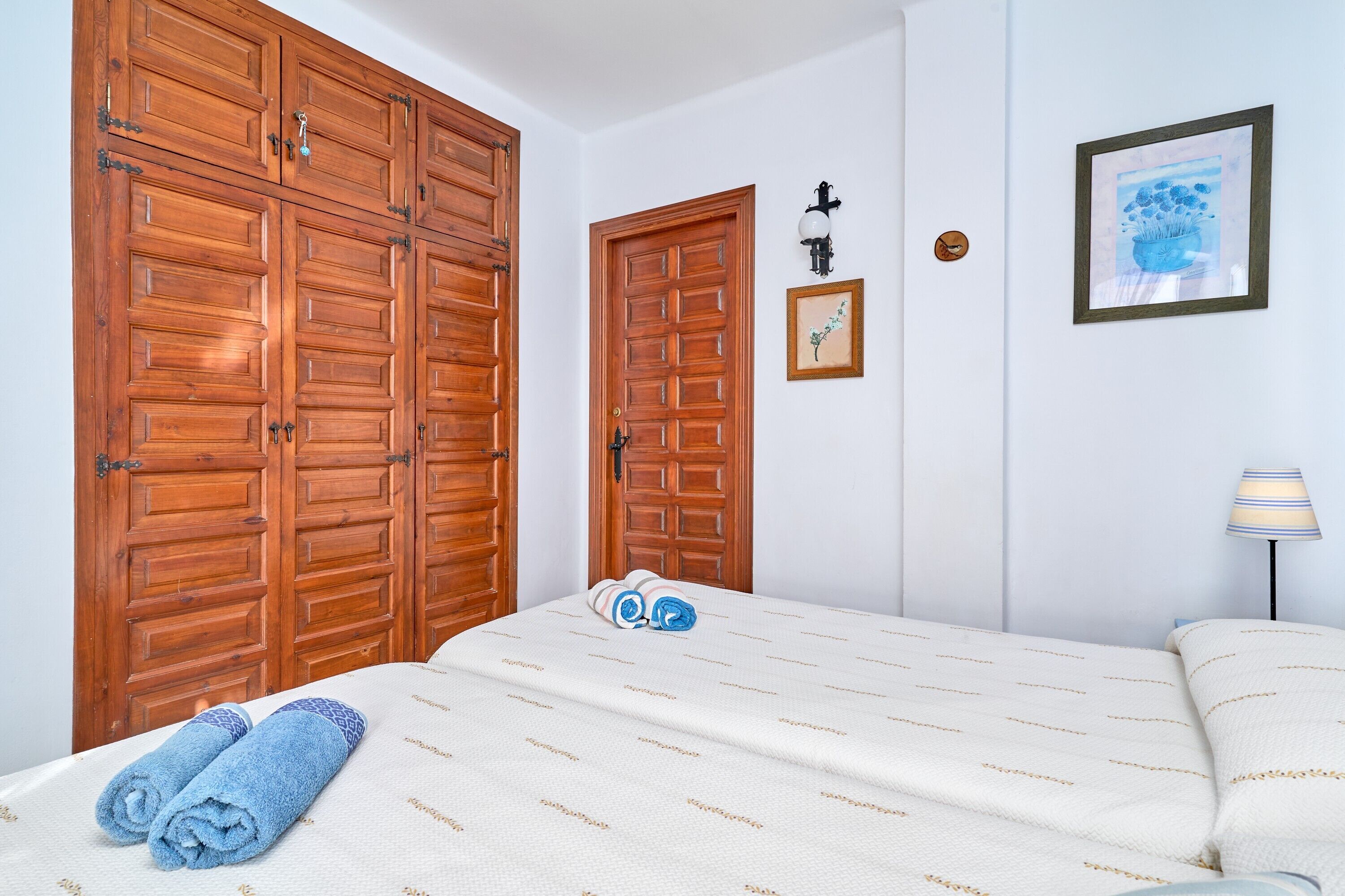 A bedroom with a bed, a wooden wardrobe, a framed picture, and a wall-mounted lamp.