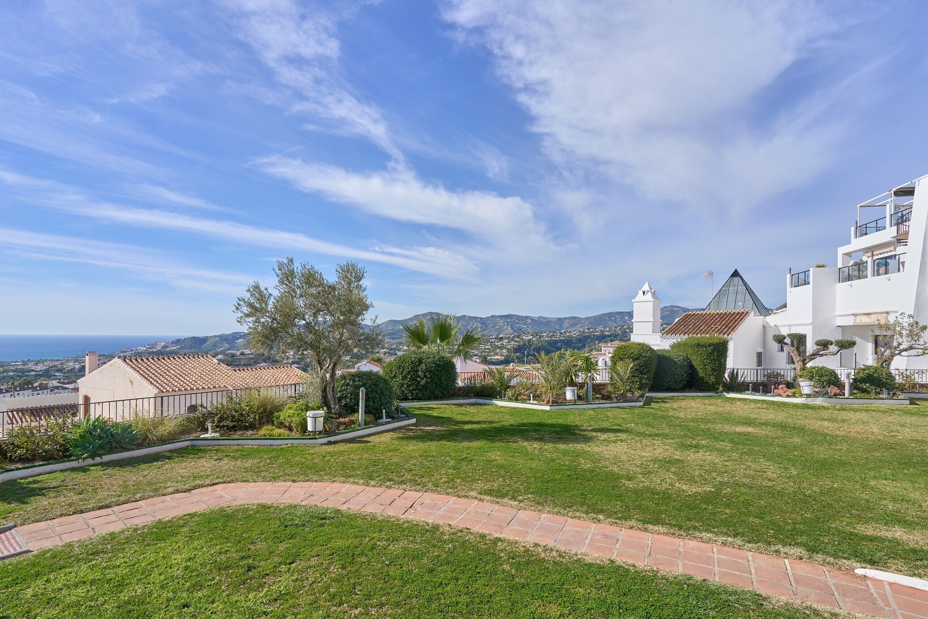 A residential area with a paved pathway, a grassy lawn, and a white building with a tiled roof.