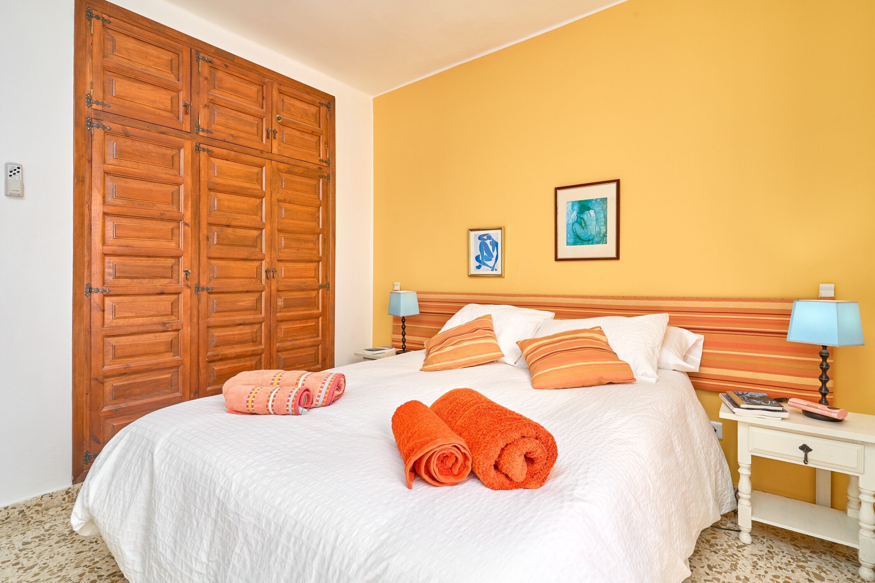 A hotel room with a large wooden wardrobe, a bed with white bedding, two pillows, a nightstand with a lamp, and two framed pictures on the wall.