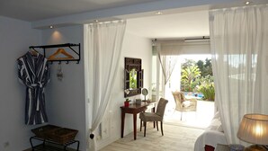 Une chambre d’hôtel comprenant un lit, un bureau avec une chaise, un fauteuil en osier, une petite table avec une lampe, et une vue sur une piscine et la verdure environnante.