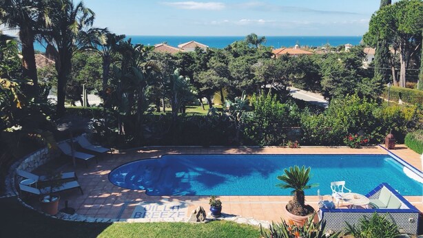 Une piscine avec vue sur l’océan, entourée de palmiers et dotée d’un espace patio avec des chaises longues.