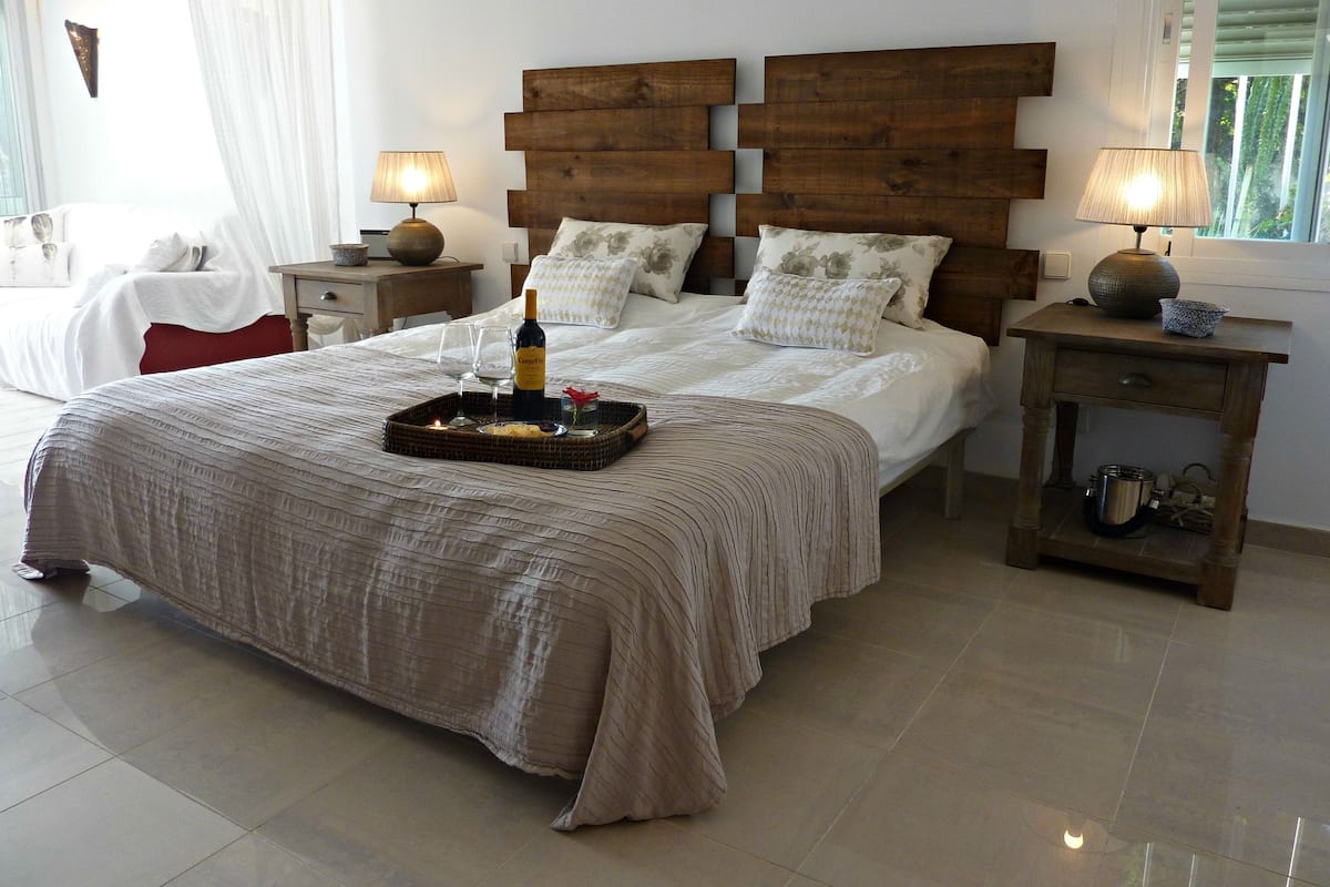 Una cama bien hecha con una bandeja de copas de vino, una botella y una vela, flanqueada por cabeceros y mesitas de noche de madera con lámparas.