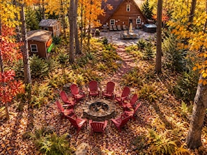 Un espacio de estar circular al aire libre con sillas rojas alrededor de una chimenea central, rodeado por un paisaje boscoso con follaje otoñal.
