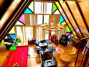 A cozy attic room with a pool table, massage chairs, a piano, and a large window with colorful panes.