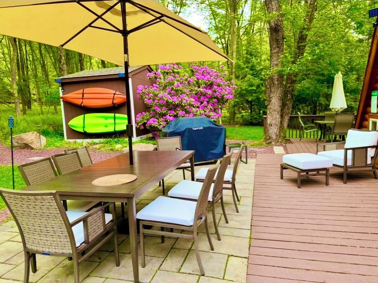 Une terrasse extérieure avec une table et des chaises en bois, un barbecue abrité et un coin jardin agrémenté de fleurs roses.