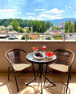 A balcony with two wicker chairs, a round table with a glass top, and a view of a cityscape with buildings and trees.