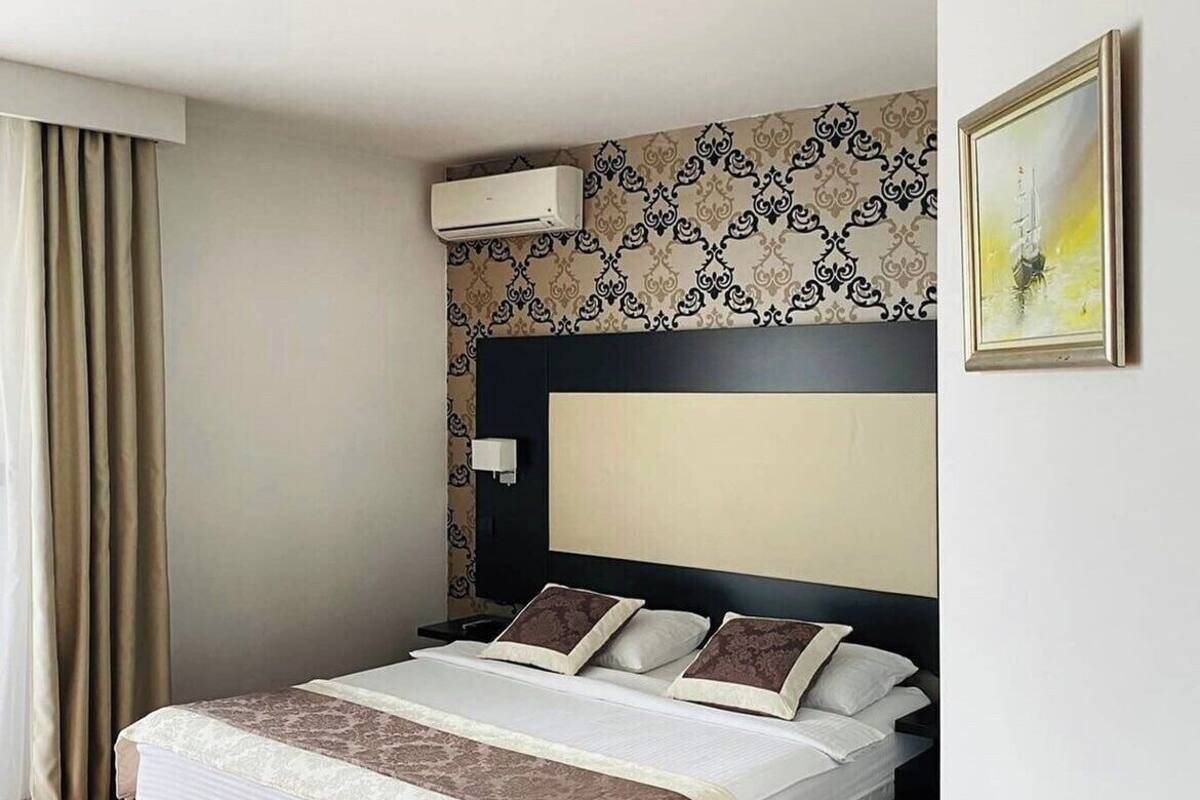 A hotel room with a large bed, an air conditioning unit, a patterned wallpaper, a painting on the wall, and a red carpet.