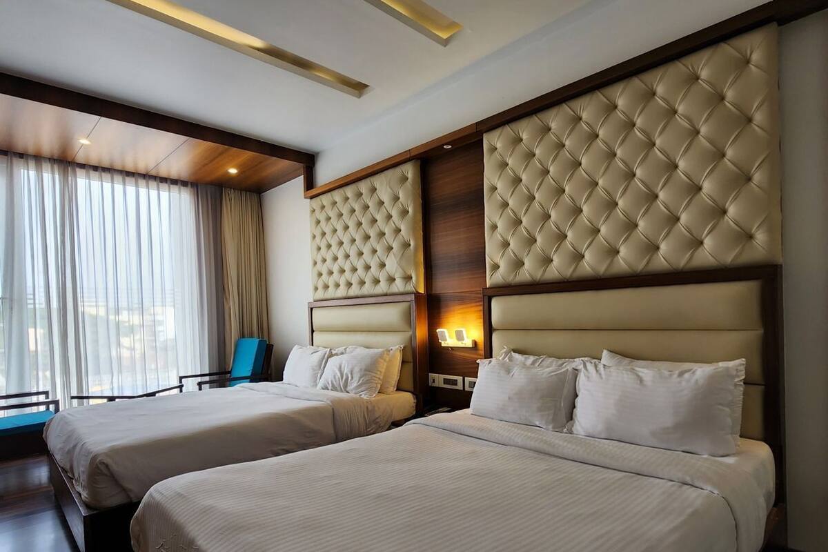 A hotel room with two beds, a large window with curtains, a wooden floor, and a modern ceiling design.