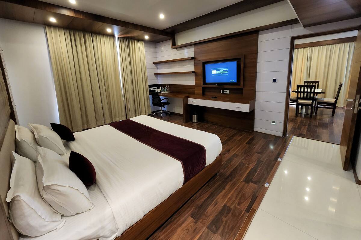 A modern hotel room with a large bed, a desk with a computer, a TV, and a dining area with chairs.