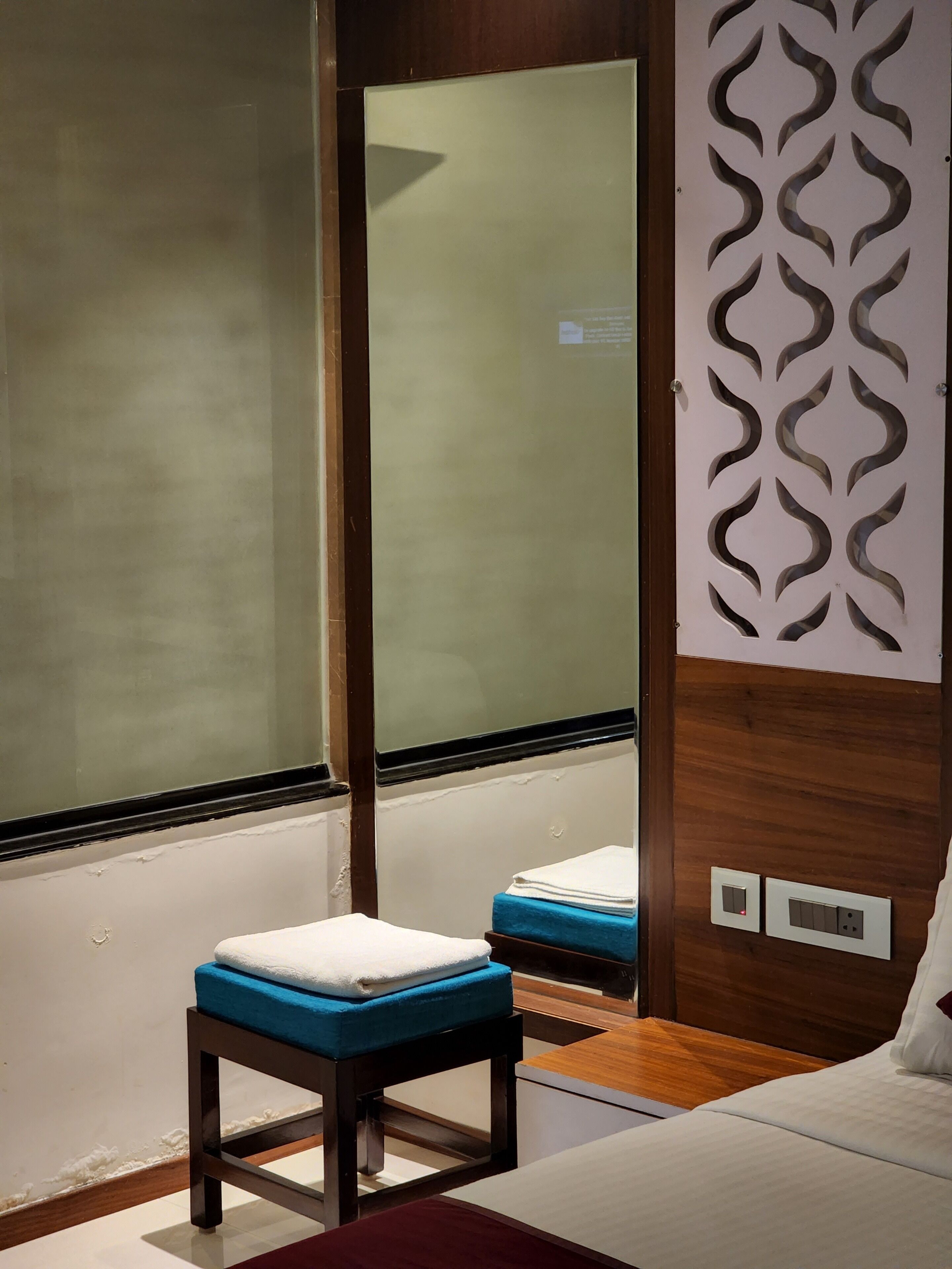 A hotel room with a bed, a small stool with a blue cushion, a wooden partition with a decorative pattern, and a window with frosted glass.
