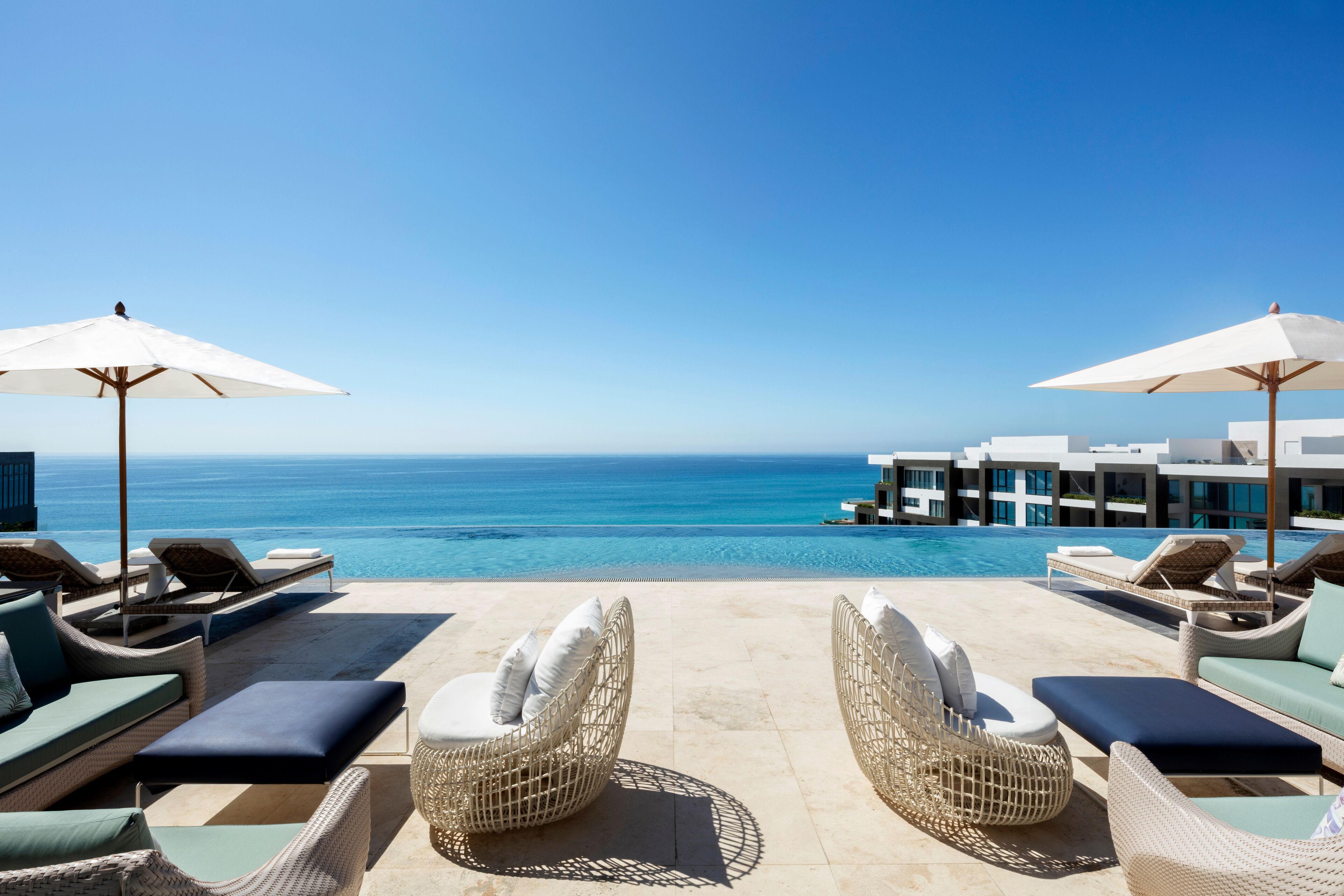 Zona junto a la piscina con sillones de mimbre y sombrillas, con vistas a un océano azul cristalino y un moderno complejo de edificios.
