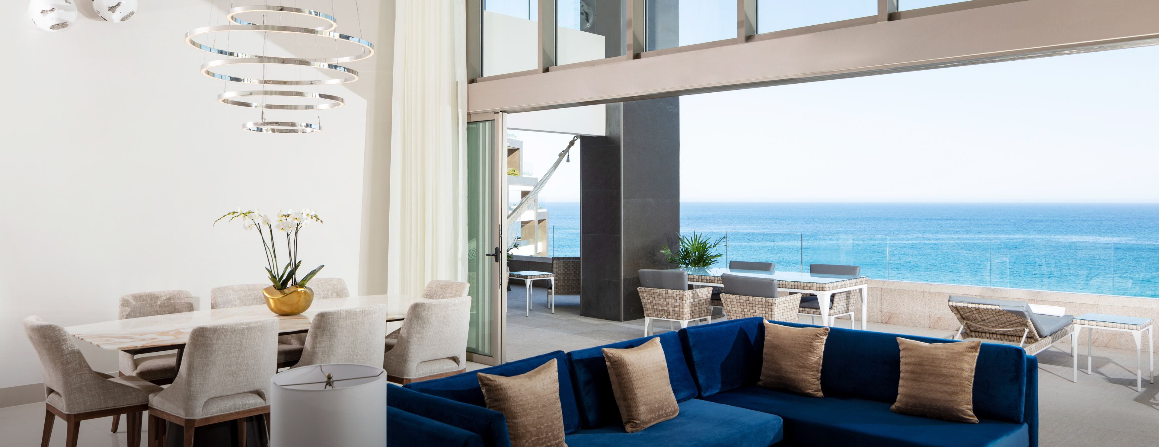 Uma sala de jantar moderna com vista para o oceano, um sofá azul e um candelabro.