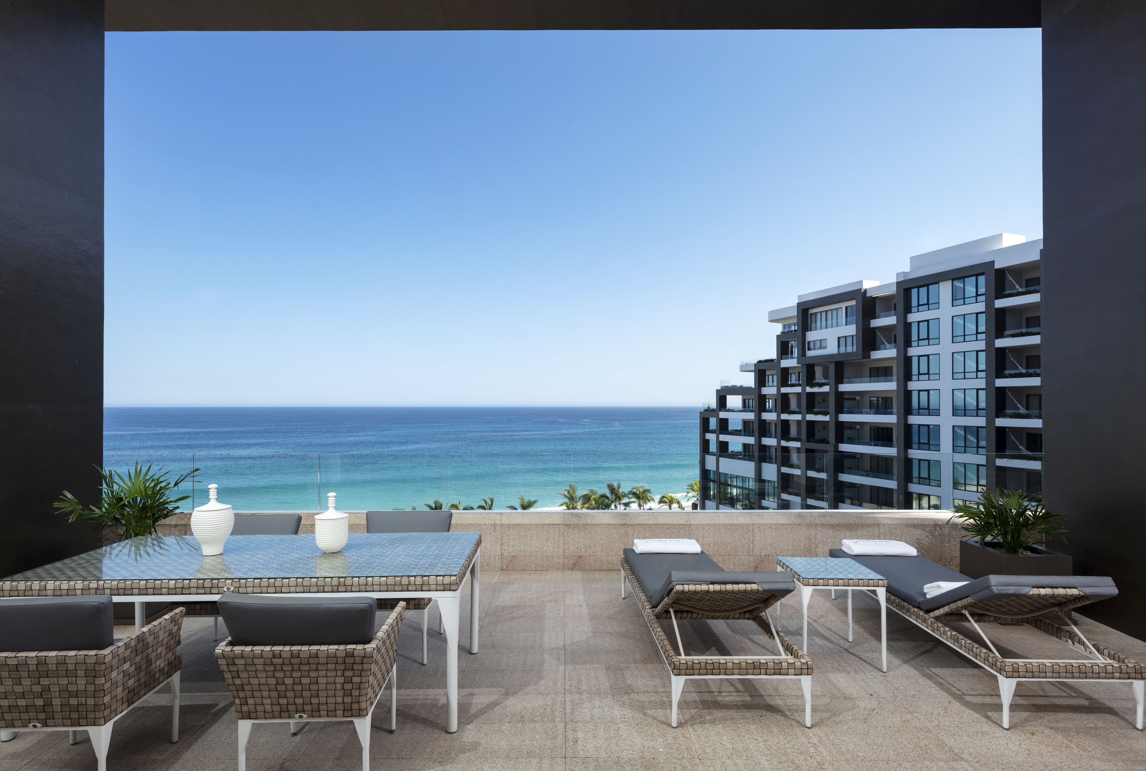 A balcony with a glass table, wicker chairs, and lounge chairs overlooking the ocean.