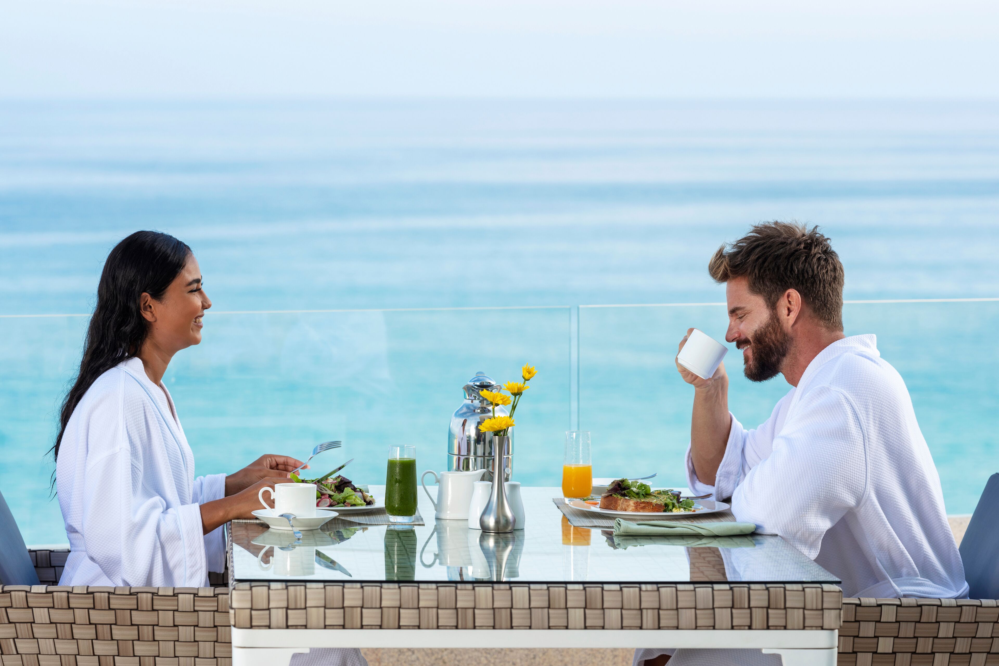 Une table de petit-déjeuner avec vue sur l’océan, une carafe, un verre de jus d’orange et une assiette de nourriture.