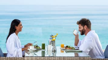 Une table de petit-déjeuner avec vue sur l’océan, une carafe, un verre de jus d’orange et une assiette de nourriture.