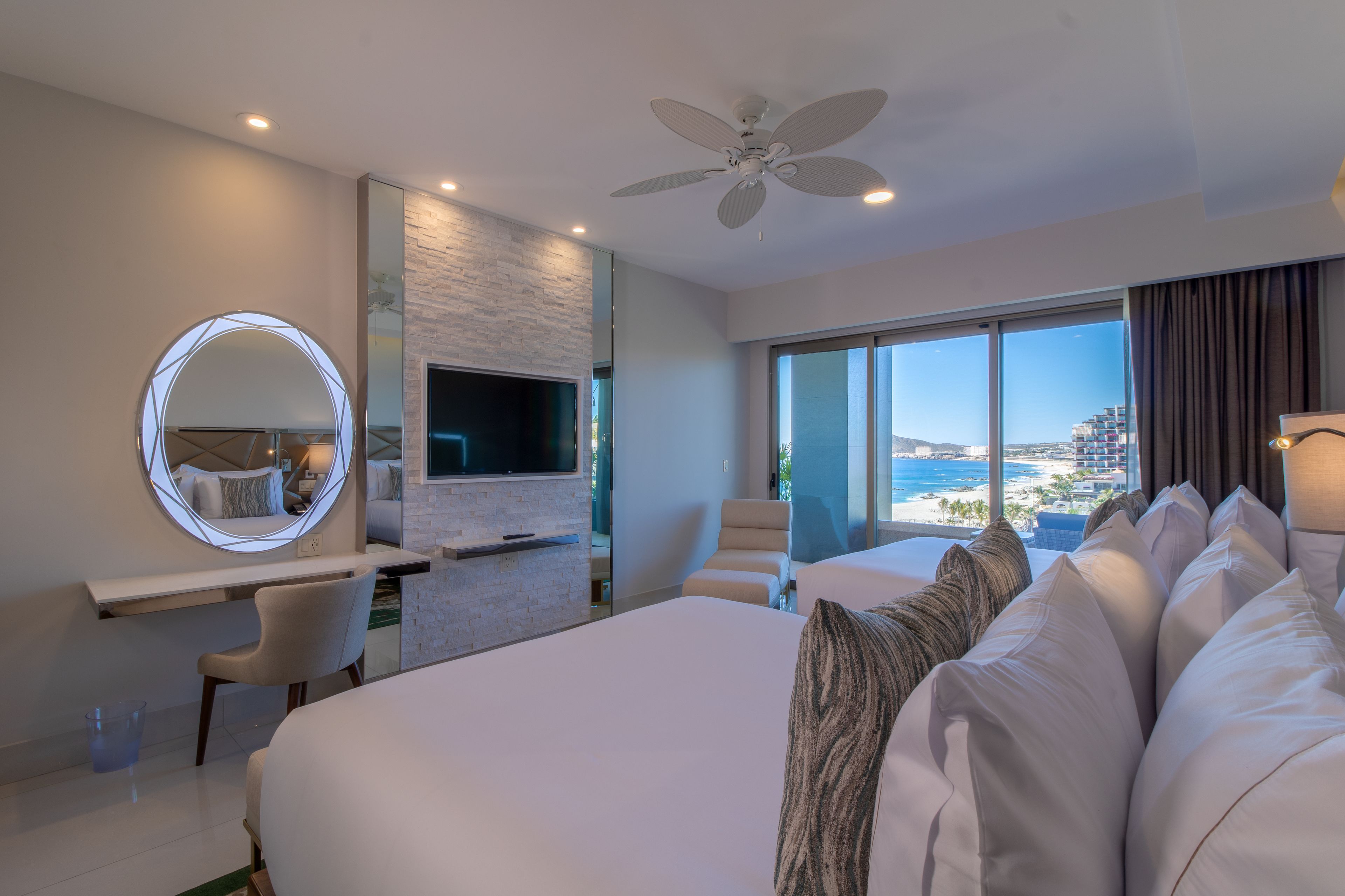 A modern hotel room with a large bed, a TV mounted on a stone wall, a round mirror, and a view of the beach.