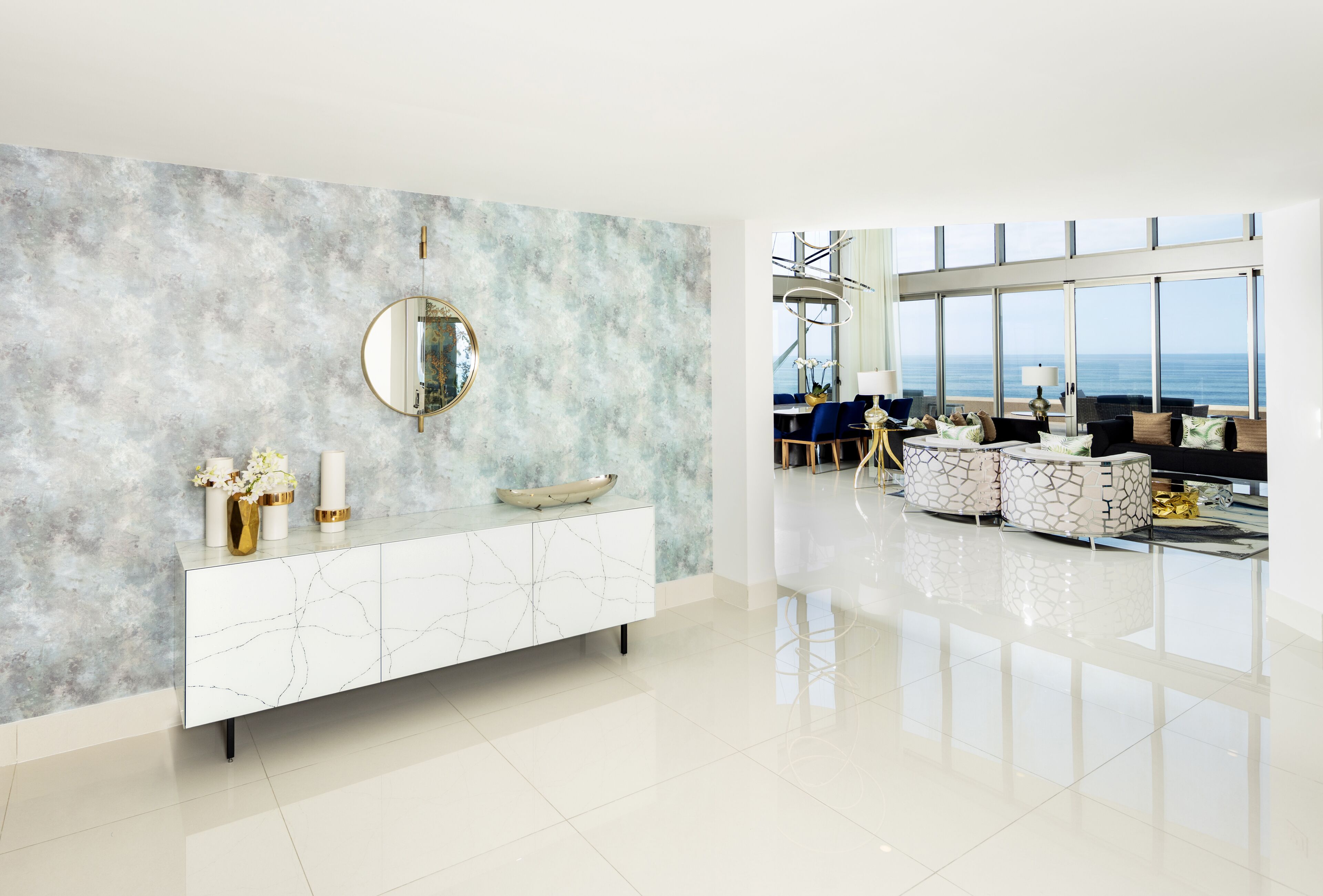 Uma sala de estar moderna com parede de mármore, um espelho redondo, um console branco e uma área de estar com vista para o mar.