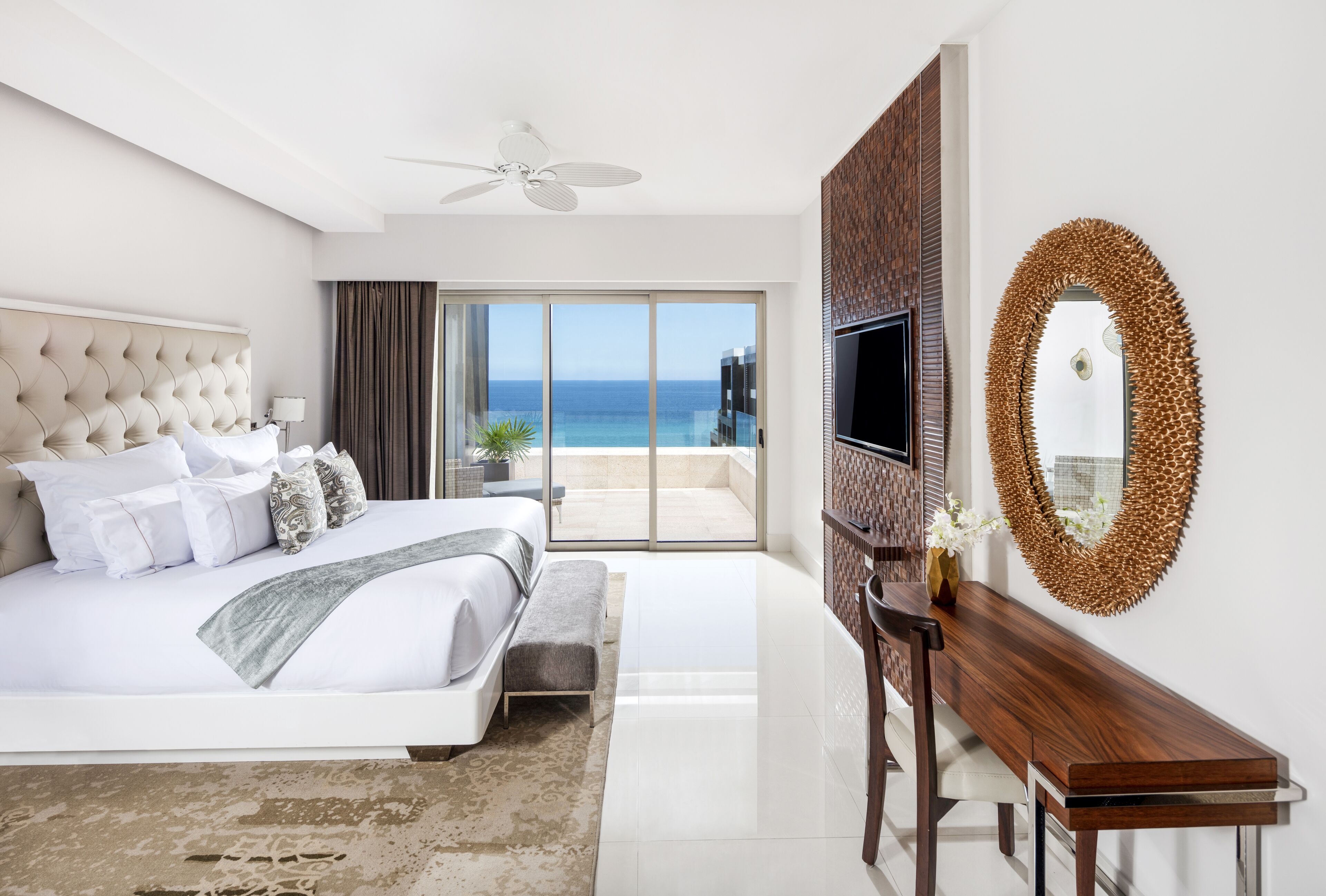 A modern hotel room with a large bed, a flat-screen TV, a wooden desk, and a balcony with a view of the ocean.