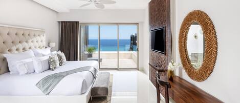 A modern hotel room with a large bed, a flat-screen TV, a wooden desk, and a balcony with a view of the ocean.