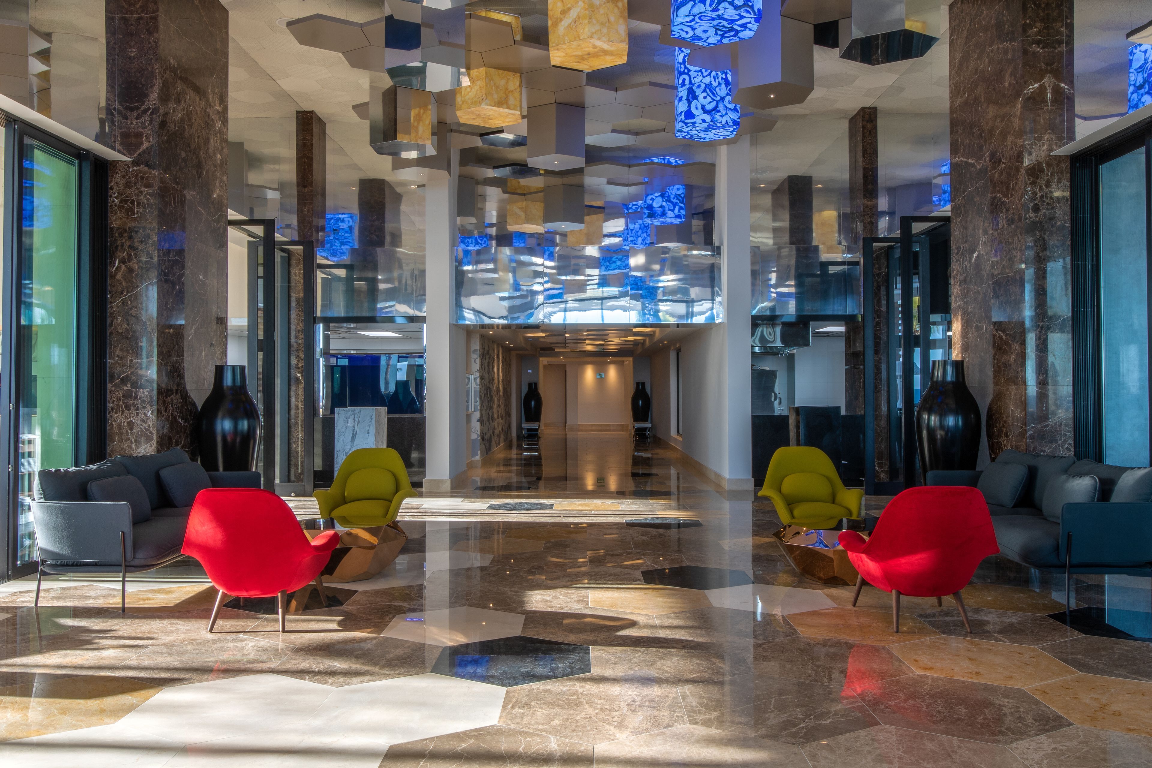 A modern lobby with a geometric floor pattern, a central staircase, and seating areas with red and yellow chairs.