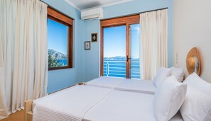 A bedroom with two beds, large windows with curtains, a view of the sea, and a straw hat on the wall. - Iris Villas Lefkada - Karavi Villa (Lefkada)