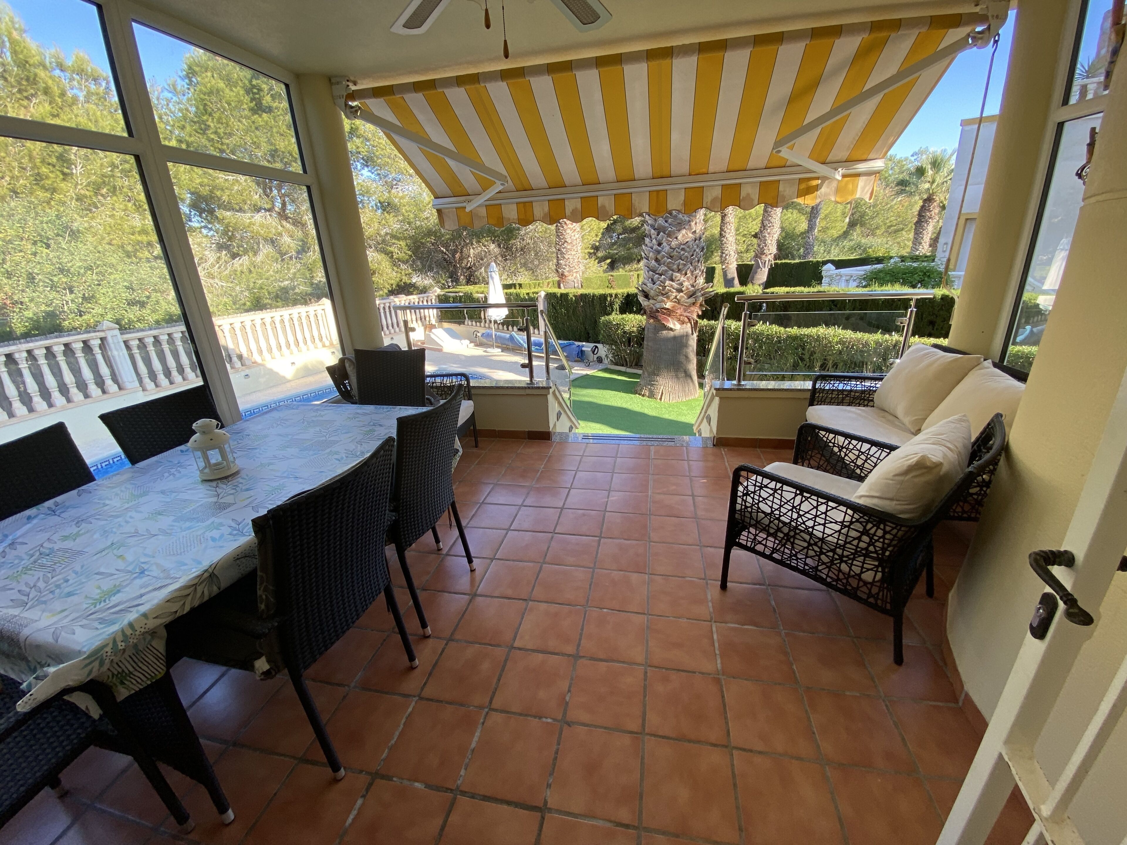 A covered patio area with a dining table and chairs, a sofa, and a view of a landscaped garden.
