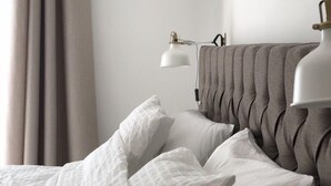 Un letto rifatto con lenzuola bianche e una testiera grigia, una lampada da comodino e una finestra con tende.