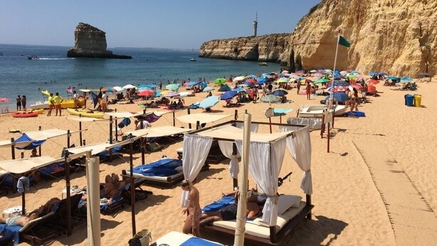 Una spiaggia con numerosi ombrelloni e tende da sole, una scogliera rocciosa e un faro sullo sfondo.