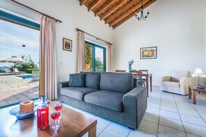A living room with a grey sofa, a wooden coffee table, and a view of a pool and beach. - Villa Ventura - Three Bedroom Villa, Sleeps 6 (Skala)
