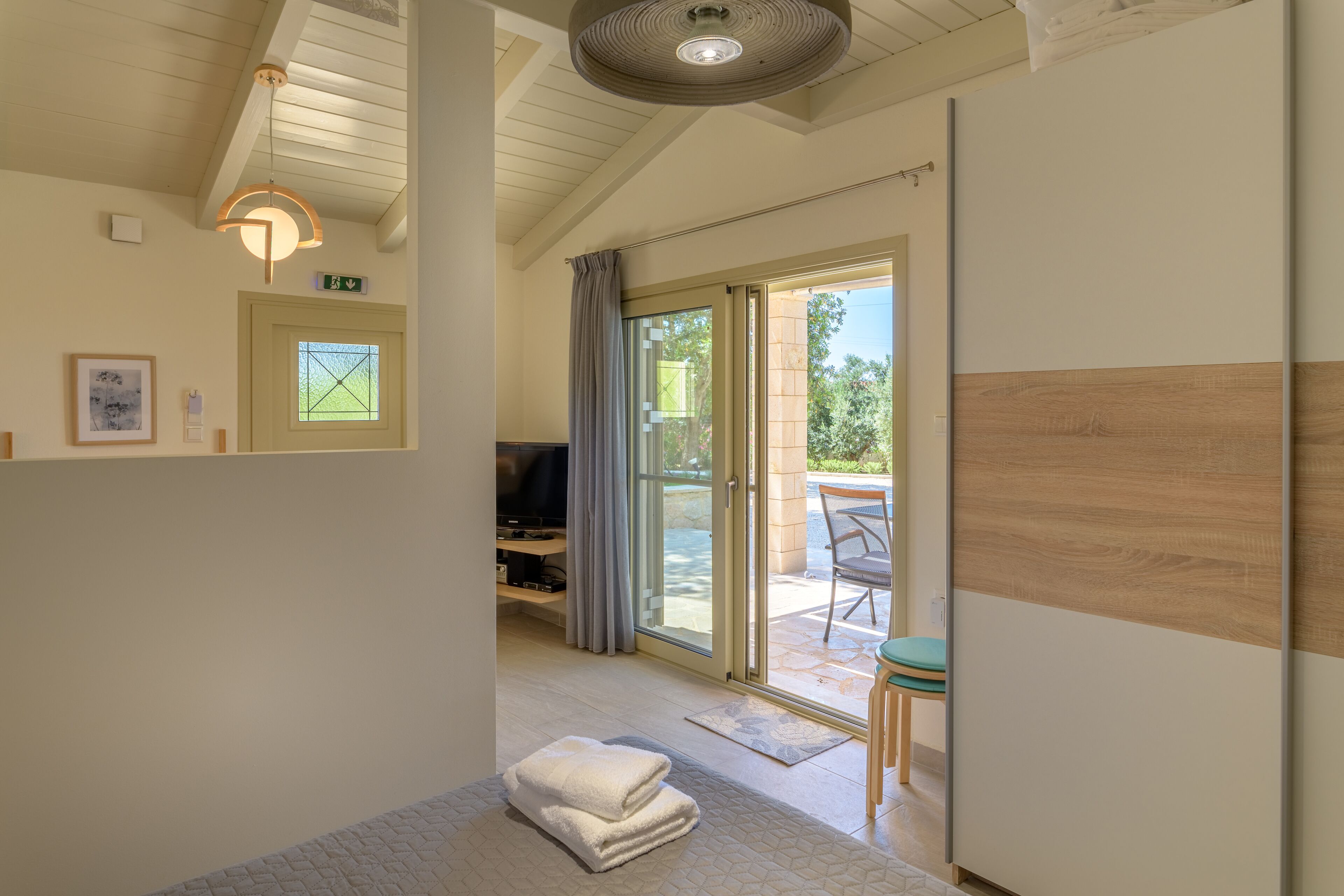 Quarto de hotel com cama, televisão, porta de vidro para uma varanda e um pequeno espaço de estar.