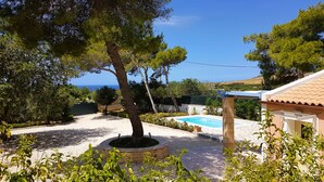 Area piscina con un pergolato, circondata da alberi e vista sul mare.