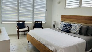 A bedroom with a bed, two chairs, a ceiling fan, and large windows with blinds. - OCEAN VIEW VILLA WITH PRIVATE POOL, STEPS FROM SECLUDED BEACH, GATED COMMUNITY! (Yabucoa)