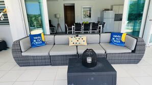 A modern living area with a grey sectional sofa, two blue pillows, a woven coffee table, and a glass door leading to a dining area. - OCEAN VIEW VILLA WITH PRIVATE POOL, STEPS FROM SECLUDED BEACH, GATED COMMUNITY! (Yabucoa)
