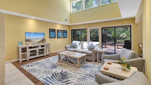 A spacious living room with a high ceiling, large windows, a sofa set, a coffee table, and a TV on a white console. - SeaPines, Ranch style home, Nicely updated, Great outdoor space, Pet friendly (hilton head)