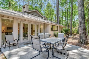 A modern outdoor patio with a dining table and chairs, surrounded by glass doors and windows, set in a wooded area. - SeaPines, Ranch style home, Nicely updated, Great outdoor space, Pet friendly (hilton head)