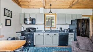 A compact kitchen with grey cabinets, a microwave, a stove, and a refrigerator. There is a dining table with chairs and a sofa in the living area. - Historic Ranger Station in the heart of Nemo, SD (Nemo)