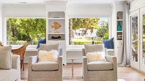 A modern living room with two grey armchairs, a white sofa, and a small wooden coffee table. There are shelves with decorative items and a view of a garden through large windows. - 63 South Sea Pines: A BESTNEST by Beverly Serral (Hilton Head Island)