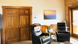 A wooden-paneled room with a large wooden door, two leather chairs, a small table with a lamp, and a painting of a sunset.