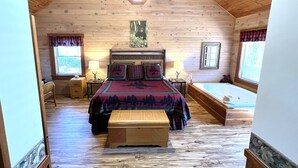 A cozy cabin-style bedroom with a wooden ceiling, a bed with a forest-themed bedspread, a wooden bench, and a ceiling fan.