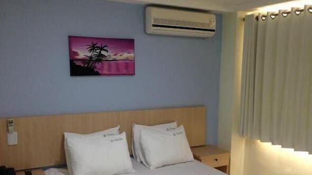 Een hotelkamer met een bed, twee nachtkastjes, een airconditioner, een schilderij en een raam met gordijnen.