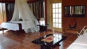 A bedroom with a four-poster bed, a bedside table with a lamp, a small table with a candle, and a large window with curtains. - Hotel Teocalli (Tepoztlán)