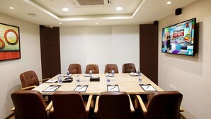 Una sala de conferencias con una larga mesa, seis sillas de cuero marrón, tarjetas de identificación, botellas de agua y un televisor mostrando '#TrendingNews'.