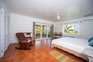 Un dormitorio con cama, una silla, ventilador de techo y vistas a palmeras a través de la ventana.