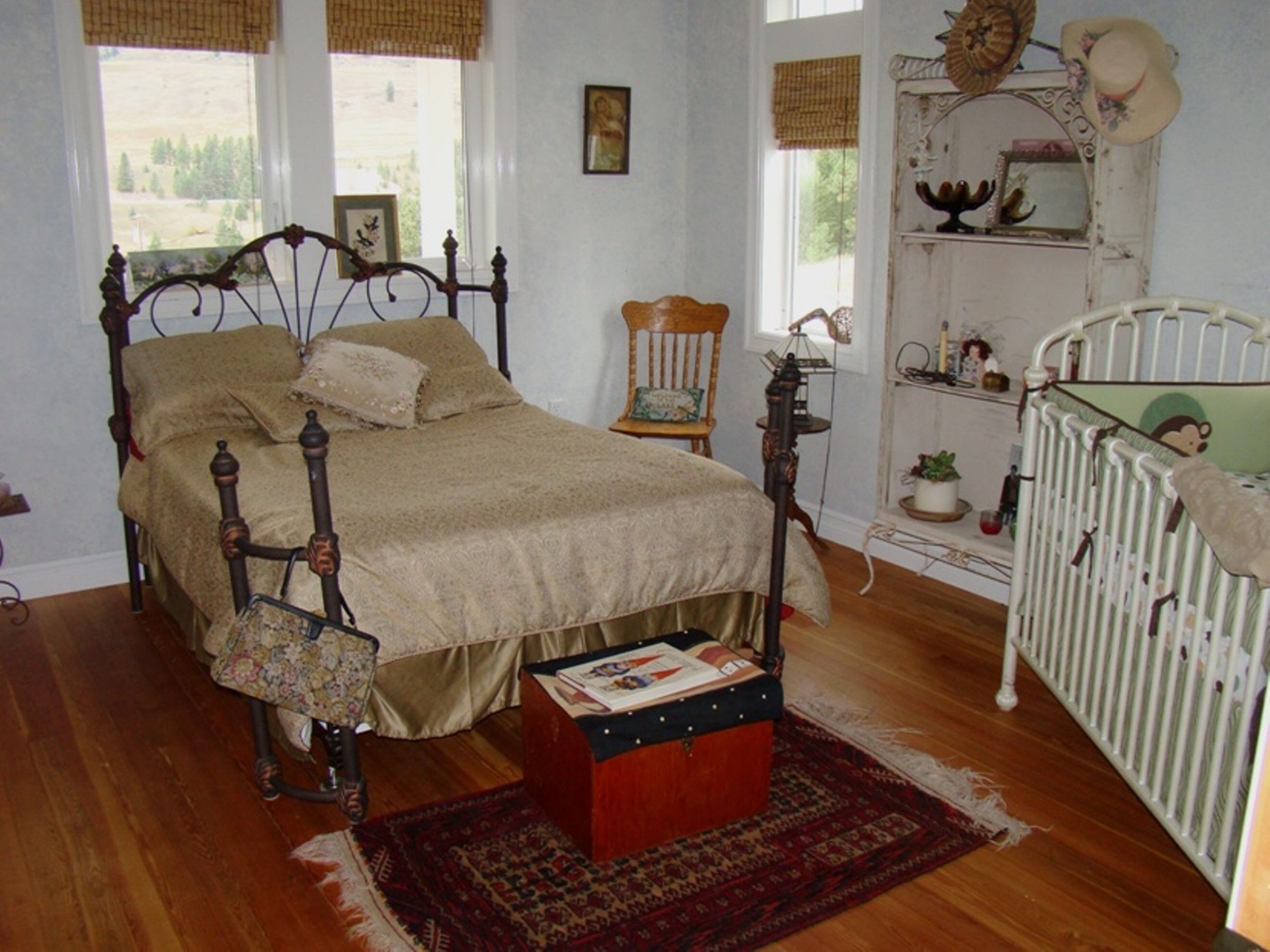 Ein Schlafzimmer mit Himmelbett, einer hölzernen Kommode, einem kleinen Stuhl, einem weißen Babybett und einem Fenster mit Blick auf Bäume.
