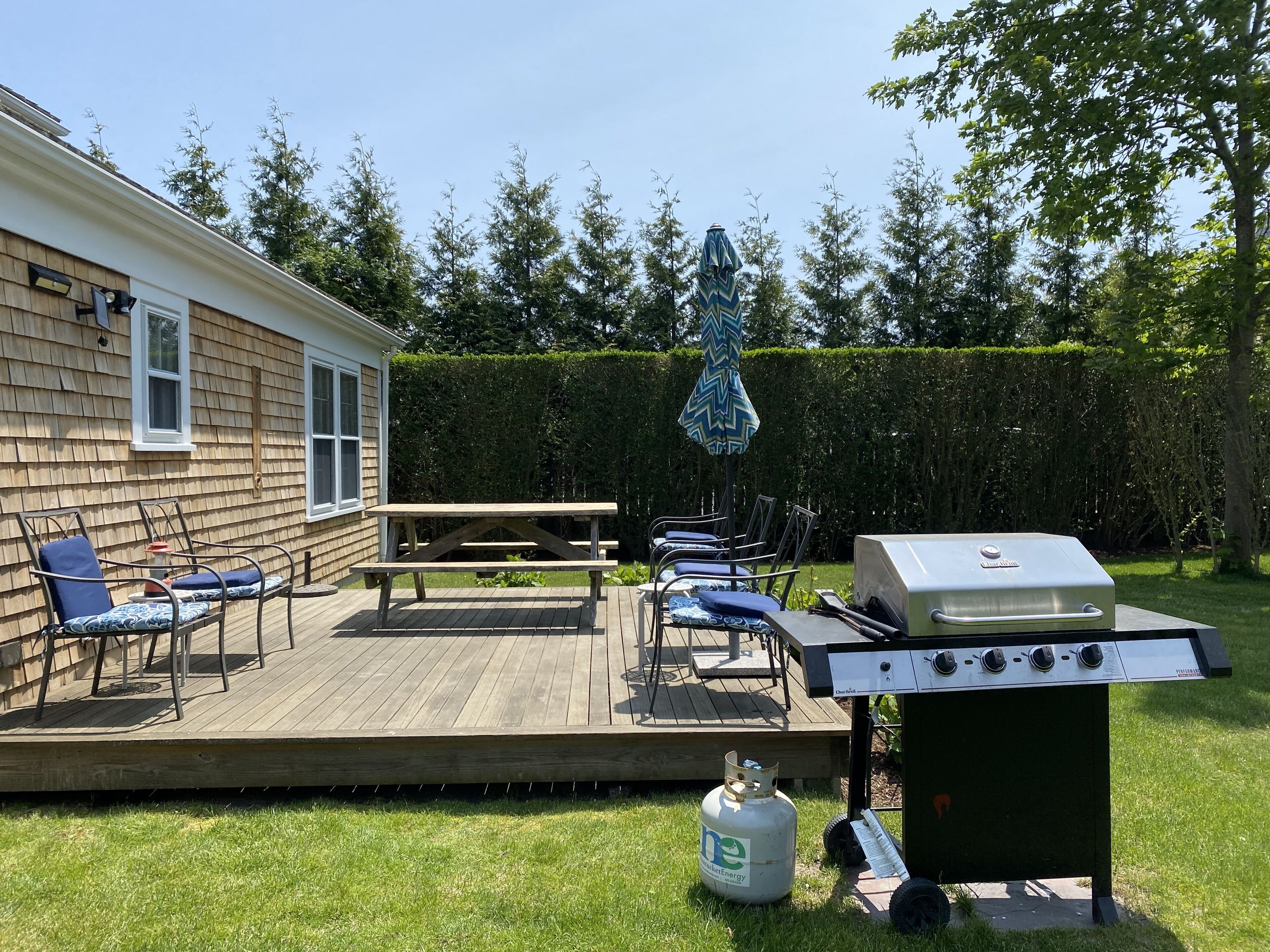 Ein Garten mit einer Holzterrasse, einem Grill und Gartenmöbeln bestehend aus Stühlen und einem Tisch.