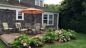 A deck with outdoor furniture and a large umbrella, surrounded by a garden with hydrangea plants. - BEAUTIFUL COTTAGE on ORANGE ST, WALK TO MAIN STREET! (Nantucket)