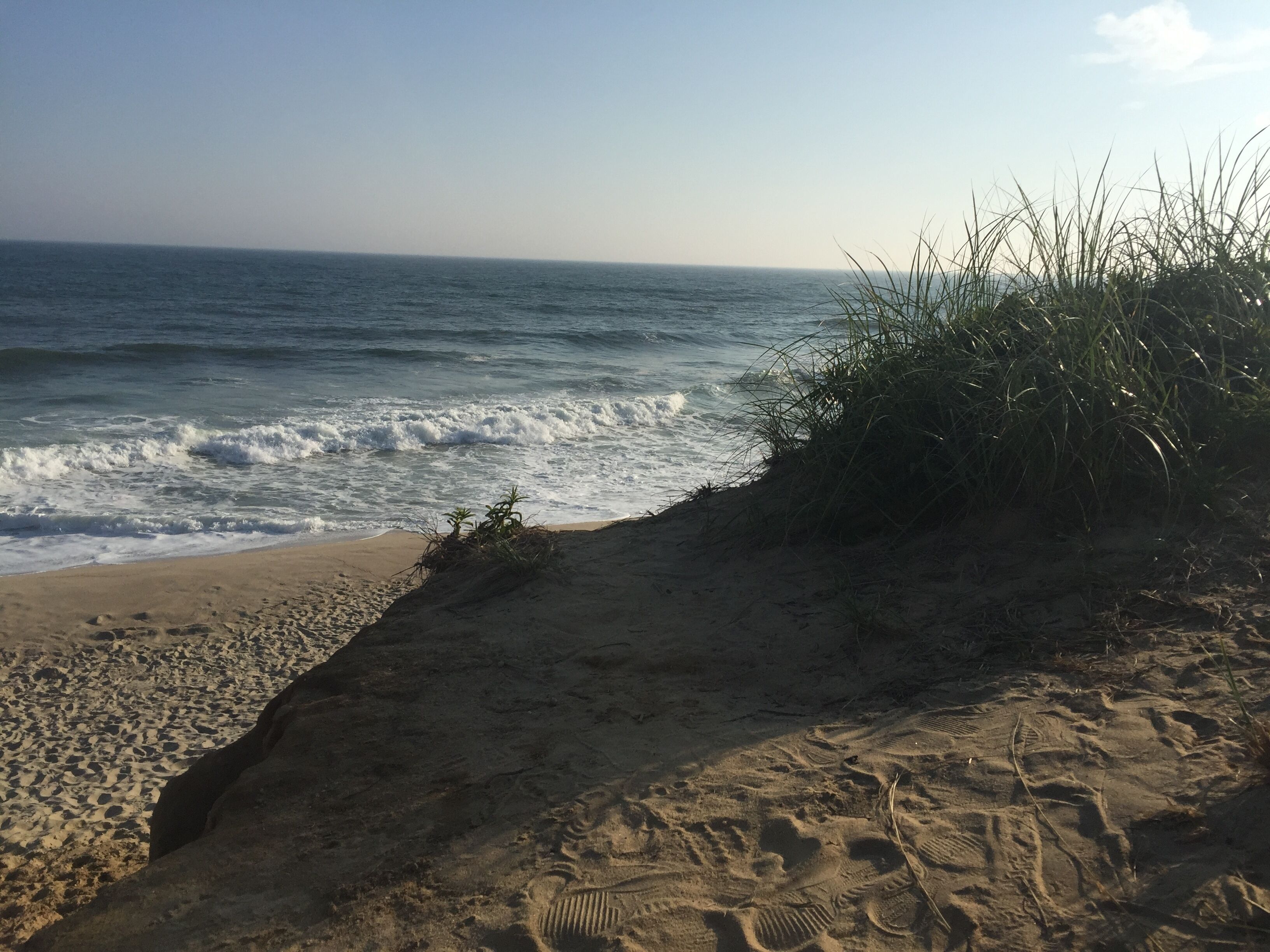 A sandy beach with footprints leading to the water, a grassy area, and waves crashing onto the shore.