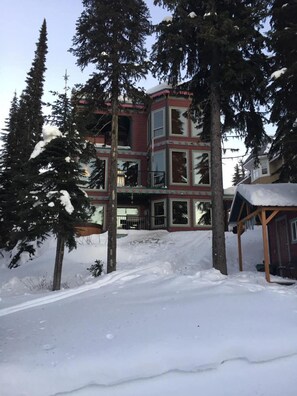 A multi-story building with large windows and a balcony, surrounded by snow-covered ground and tall evergreen trees.